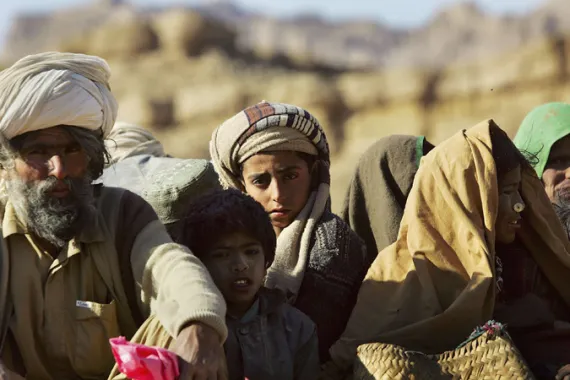 یک گروه از مردم بلوچ، شامل مردان، زنان و کودکان، با لباس‌های سنتی و پوشش‌های محلی، در فضای باز و منطقه‌ای کوهستانی و صحرایی نشسته‌اند [منبع تصویر: الجزیره]